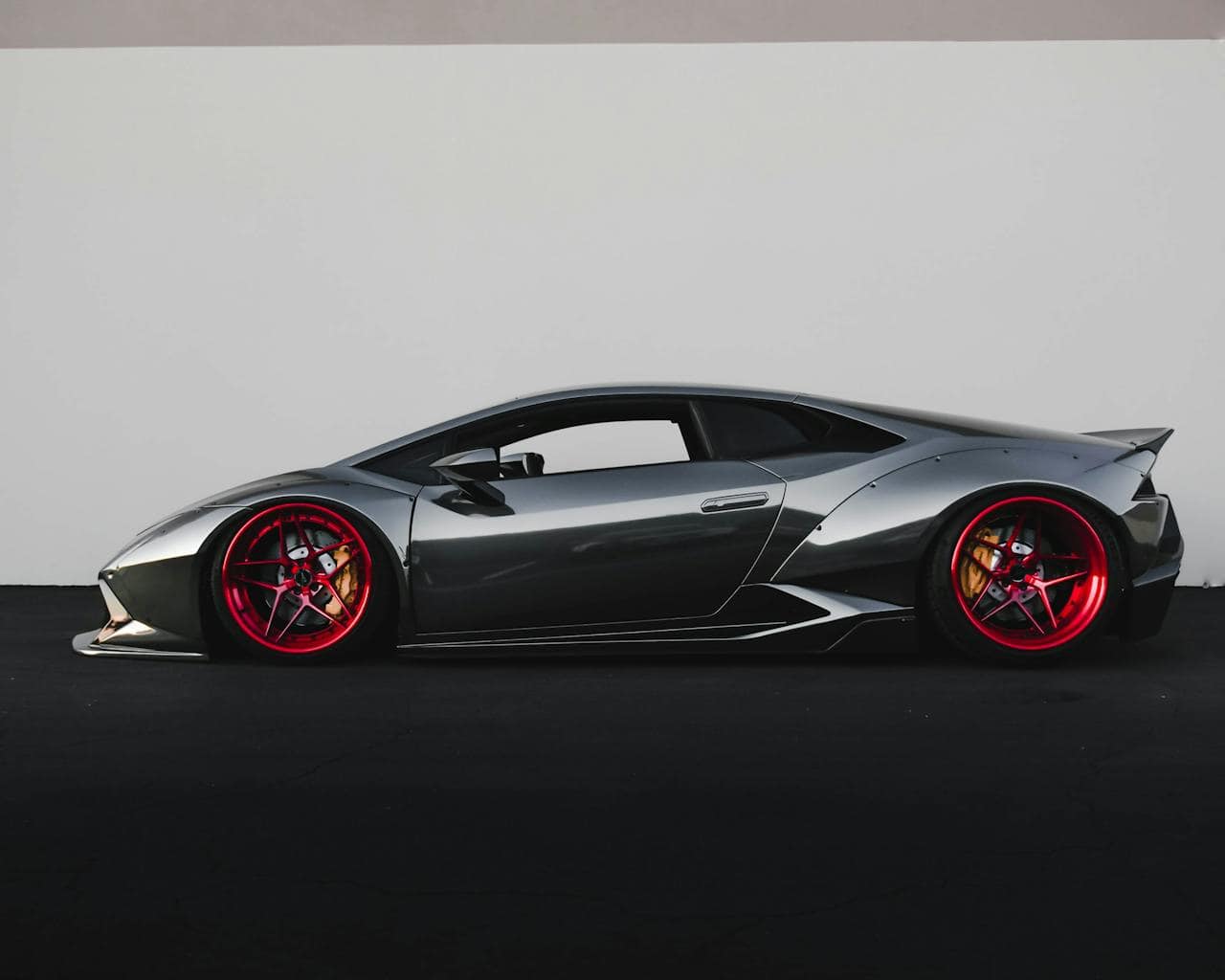 A sleek, metallic gray sports car with bright red rims is parked on a dark surface against a plain, light-colored wall. The custom auto shop in Oakland County MI has enhanced its low, aerodynamic profile and modern design features to perfection.