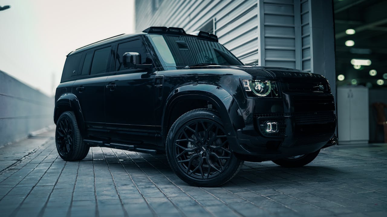 A sleek, black SUV with tinted windows parked on a cobblestone surface showcases its modern, angular design. Nearby, a metal-paneled building hints at its origin—a custom auto shop in Oakland County, MI—and offers a partial view of another structure.