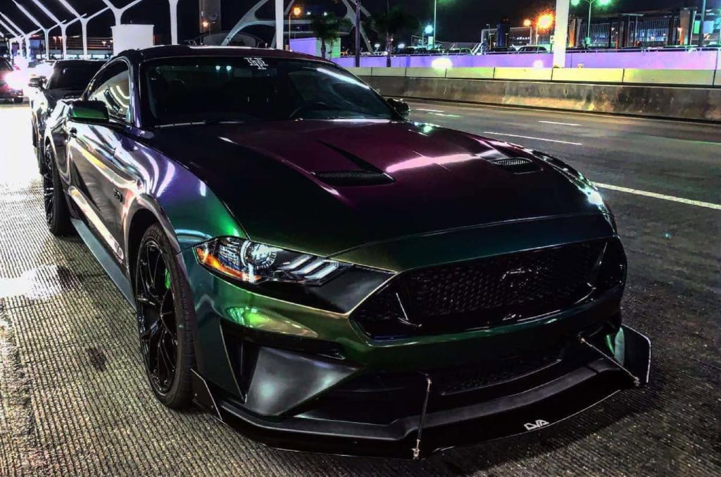 A sleek, metallic green sports car with black accents, crafted by a custom auto shop in Oakland County MI, sits parked on a city street at night. Its modern design, featuring a prominent grille and sharp headlights, gleams under the bright streetlights.