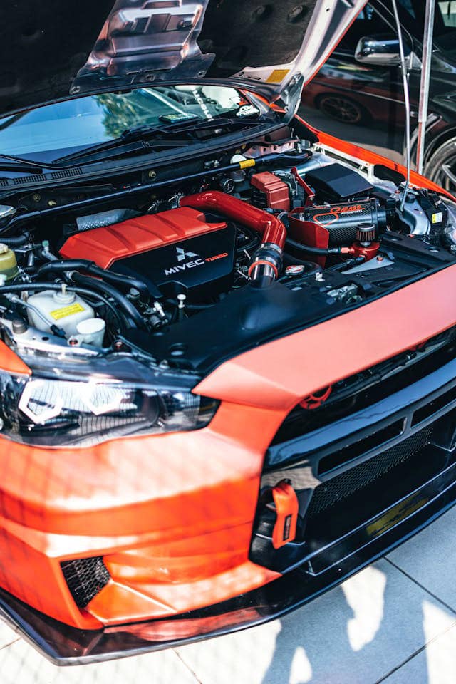 Close-up of a red Mitsubishi with its hood open, showcasing an engine labeled "MIVEC Turbo" and striking red components like the intake pipe and strut bar. Captured in daylight, this custom beauty from an auto shop in Oakland County, MI, stands parked outdoors.