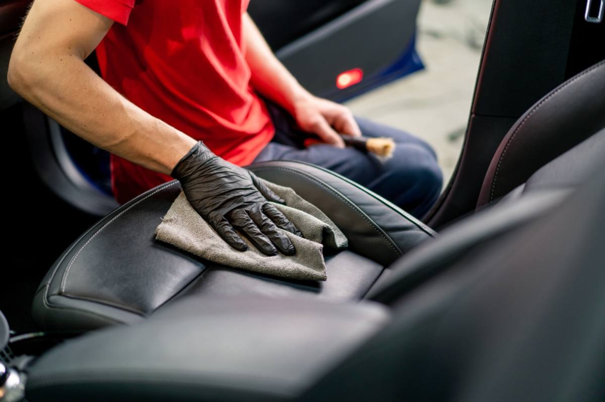 A person wearing black gloves cleans a car’s black leather seat with a gray cloth. The individual, dressed in a red shirt, is inside the vehicle, focusing on the seat’s surface.