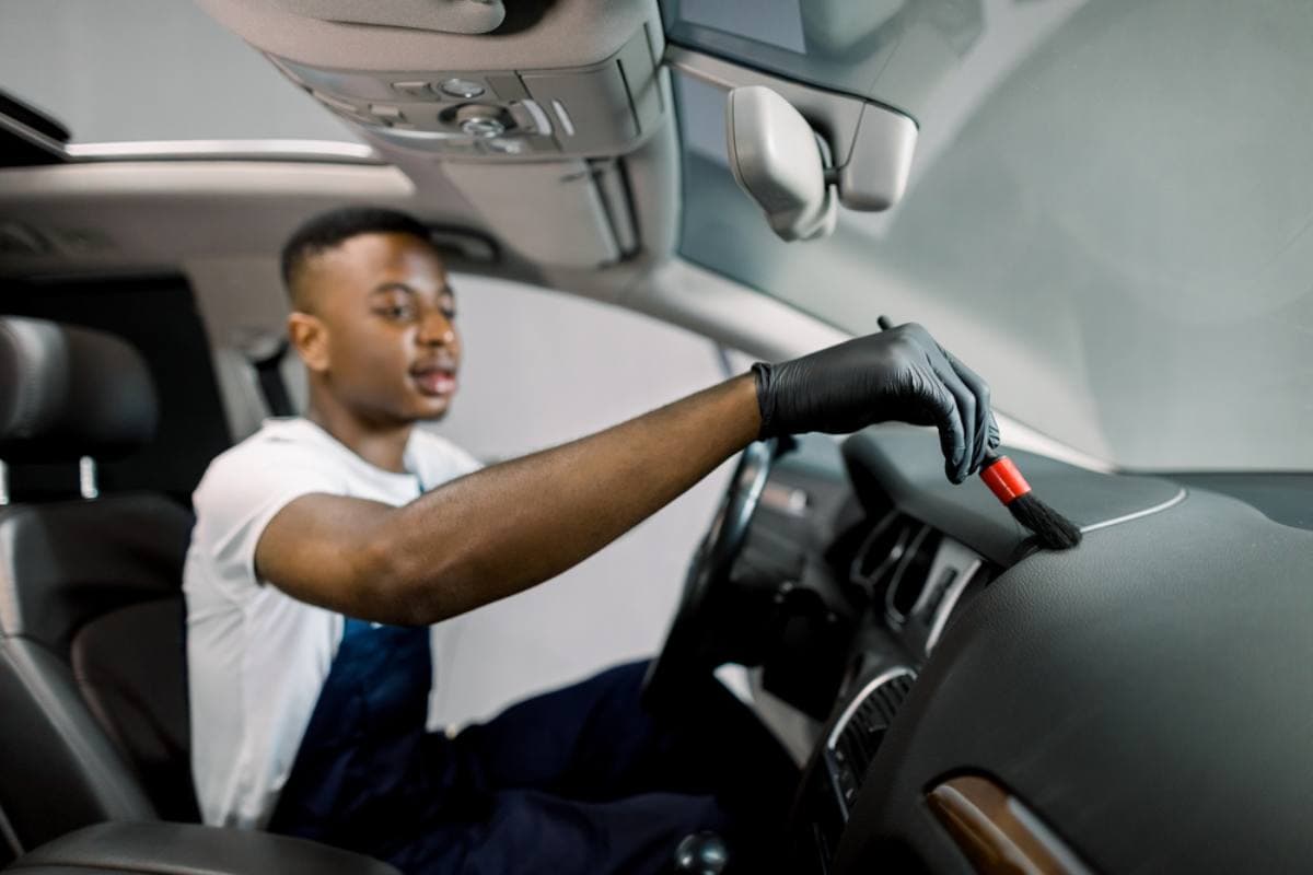 A person wearing black gloves uses a small brush to clean the dashboard of a car interior, focusing on detailing near the windshield.