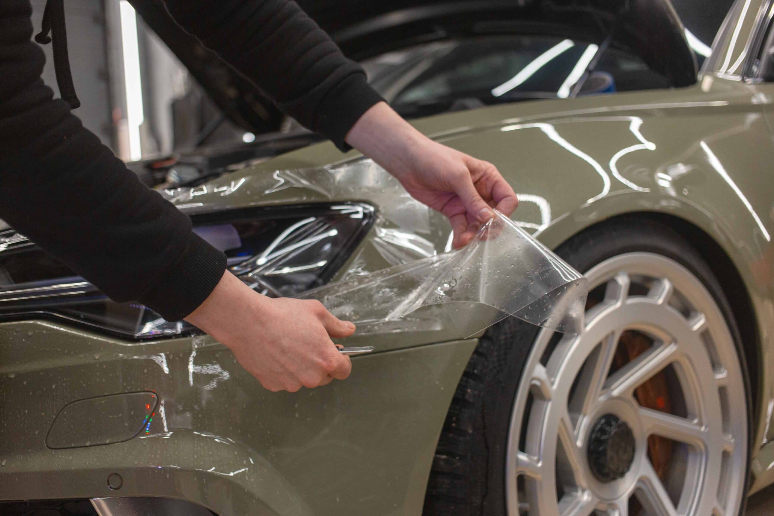 At a custom auto shop in Oakland County, MI, a person meticulously applies a clear protective film to the front fender of a matte olive green car, concentrating on the area above the wheel. The hood is open, revealing their black-sleeved arm as they work with precision.