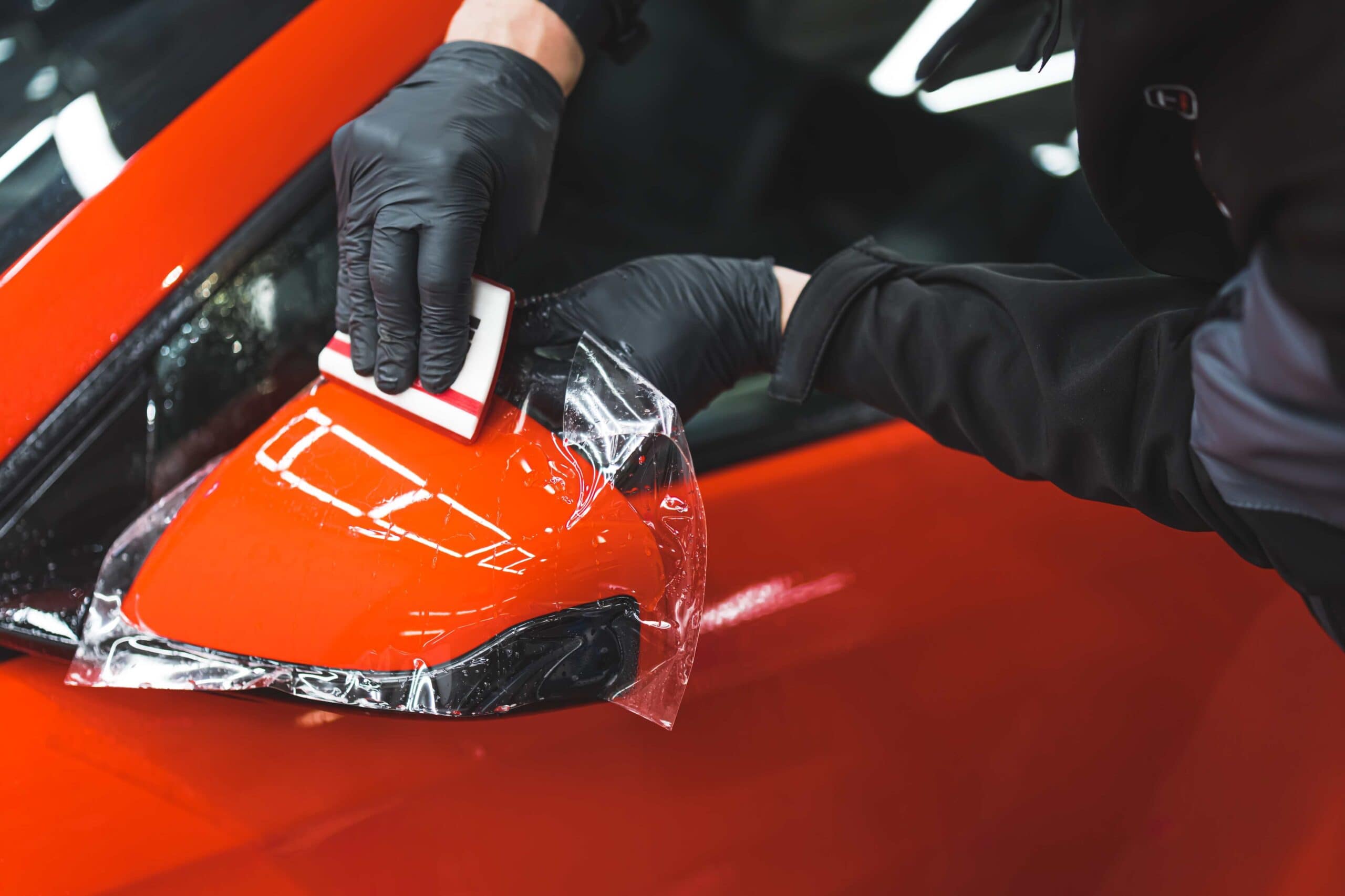 At a custom auto shop in Oakland County, MI, someone wearing black gloves expertly applies a protective film to a red car's side mirror using a small tool, ensuring the smoothest application process.