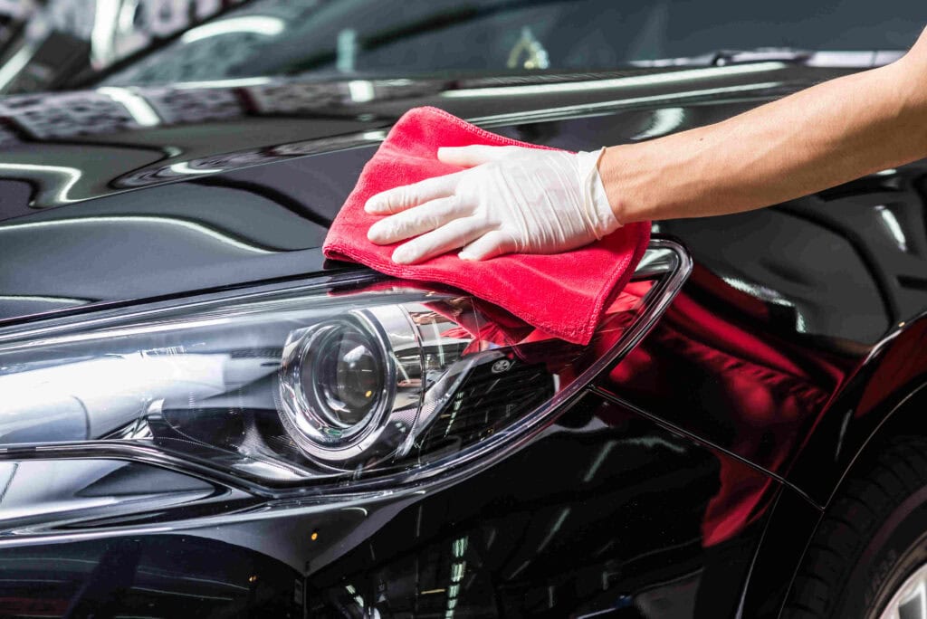 At a custom auto shop in Oakland County, MI, a person wearing a white glove uses a red cloth to polish the shiny hood of a black car near the left headlight. Reflections of lights dance across the car's pristine surface.