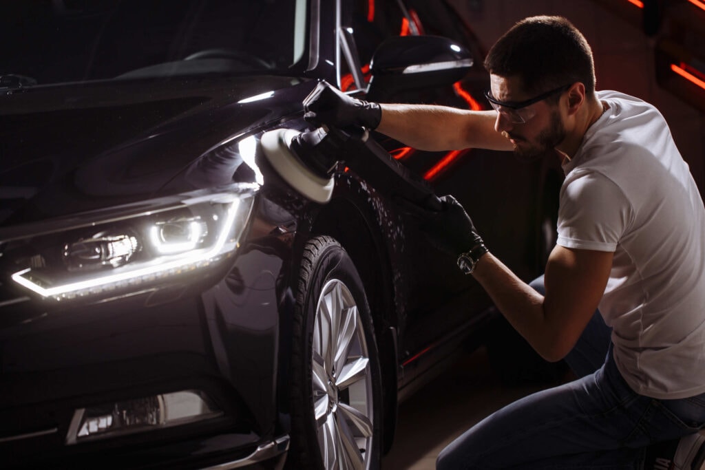 In a custom auto shop in Oakland County, MI, a person wearing gloves and safety glasses expertly uses a buffer to polish the hood of a sleek black car. The car's headlights illuminate the red light reflections on its surface, adding a vibrant glow to the indoor scene.