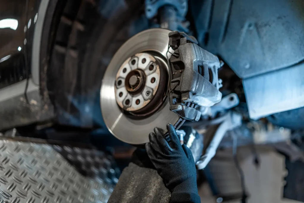 A mechanic wearing black gloves inspects or repairs a car’s disc brake system, focusing on the brake caliper and rotor inside an automotive workshop.