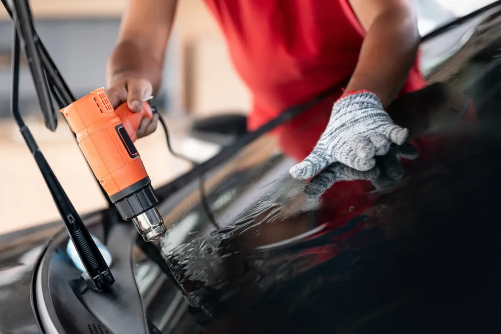 A person wearing a red shirt and a protective glove uses a heat gun to apply or remove a film from a car windshield.